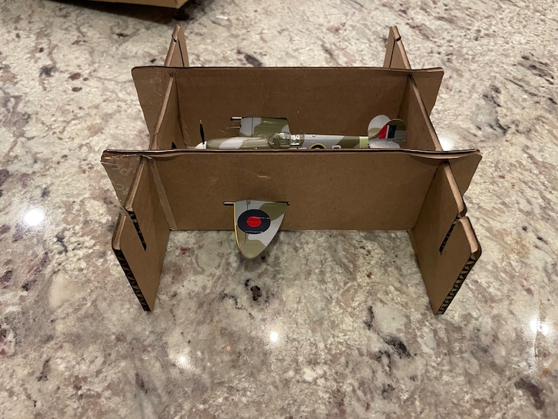 May include: A cardboard box with a model airplane inside. The airplane is painted in camouflage colors with a red and white roundel on the tail.