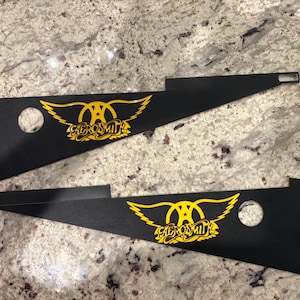 Op de afbeelding: Twee zwarte metalen panelen met een gele Aerosmith-logo. Elk paneel heeft een cirkelvormig gat en een rechthoekige uitsparing. Het logo heeft een gestileerd vleugelontwerp en de naam van de band.