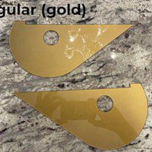 May include: Two gold-colored, flat, teardrop-shaped pieces with a smooth, reflective surface. Each piece features a circular cutout near the center and a small notch along the top edge. The text "gular (gold)" is visible at the top left corner.