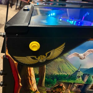 Puede incluir: Una máquina de pinball negra con un botón amarillo y un emblema dorado. La obra de arte presenta un búho nival, un paisaje montañoso y la letra "H". La máquina tiene un émbolo negro y una tapa de cristal transparente.