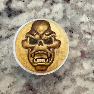 May include: A round, gold-colored button with a detailed, embossed skull design. The skull has a menacing expression with sharp teeth and deep eye sockets. The button is likely made of metal and has a white border.