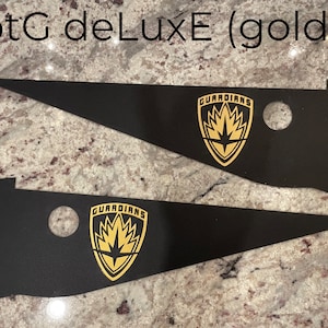Op de afbeelding: Twee zwarte vlaggen met een driehoekige vorm met gouden Guardians of the Galaxy-logo's. De logo's hebben een ster in het midden van een vlamvormig ontwerp.