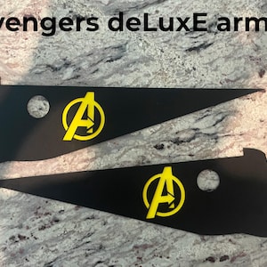 Op de afbeelding: Twee zwarte, driehoekige stukken pantser met een geel Avengers-logo. De tekst "Avengers deLuxE armor" staat bovenaan de afbeelding.