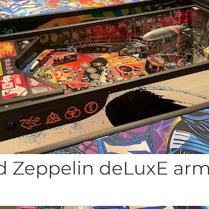 May include: A black pinball machine with a glass top. The pinball machine has a red and black playfield with a Led Zeppelin design. The machine has a black cabinet with a white and black design. The cabinet has the words "Zoso" and the Led Zeppelin symbol on it.