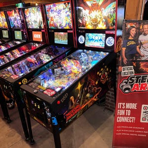 May include: A row of pinball machines with various themes, including Dungeons & Dragons, The Tyrant's Eye, and a machine with a colorful, abstract design. The machines are in a dimly lit room with brick walls and a sign that reads "It's more fun to connect!"