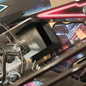 May include: Close-up of a pinball machine's inner workings, showing a metal flipper arm with a red and black painted design. The arm is attached to a metal frame with screws.