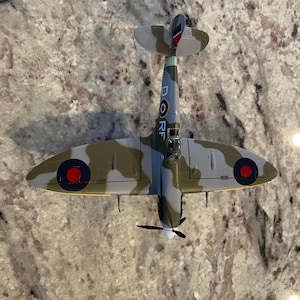 May include: A model of a Supermarine Spitfire fighter plane in camouflage paint with red, white, and blue markings. The plane has a yellow stripe on the fuselage and the letters "D" and "RF" on the side.