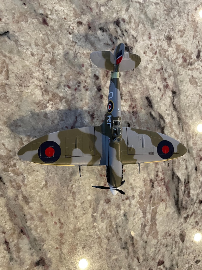 May include: A model of a Supermarine Spitfire fighter plane in camouflage paint with red, white, and blue markings. The plane has a yellow stripe on the fuselage and the letters "D" and "RF" on the side.