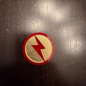 Pode incluir: Um alfinete redondo, vermelho e dourado com um design de raio vermelho. O alfinete tem um centro dourado texturizado e uma borda vermelha. Sobre uma superfície de madeira castanha escura.