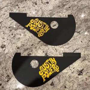 Puede incluir: Dos piezas de plástico negras y curvas con un logotipo amarillo "Austin Powers". Las piezas tienen un agujero en el centro.