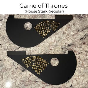 May include: Two black, triangular-shaped wall decals with a gold outline of a direwolf, the sigil of House Stark from the Game of Thrones television series. The text "Game of Thrones (House Stark)(regular)" is printed above the decals.