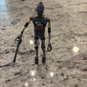 以下が含まれることがあります： スターウォーズのIG-11アクションフィギュア。中にはベビーヨーダのフィギュアが入っています。IG-11のフィギュアは主にグレーで、黄色と赤のアクセントがあります。ブラスターライフルを持っています。ベビーヨーダのフィギュアは緑と茶色です。