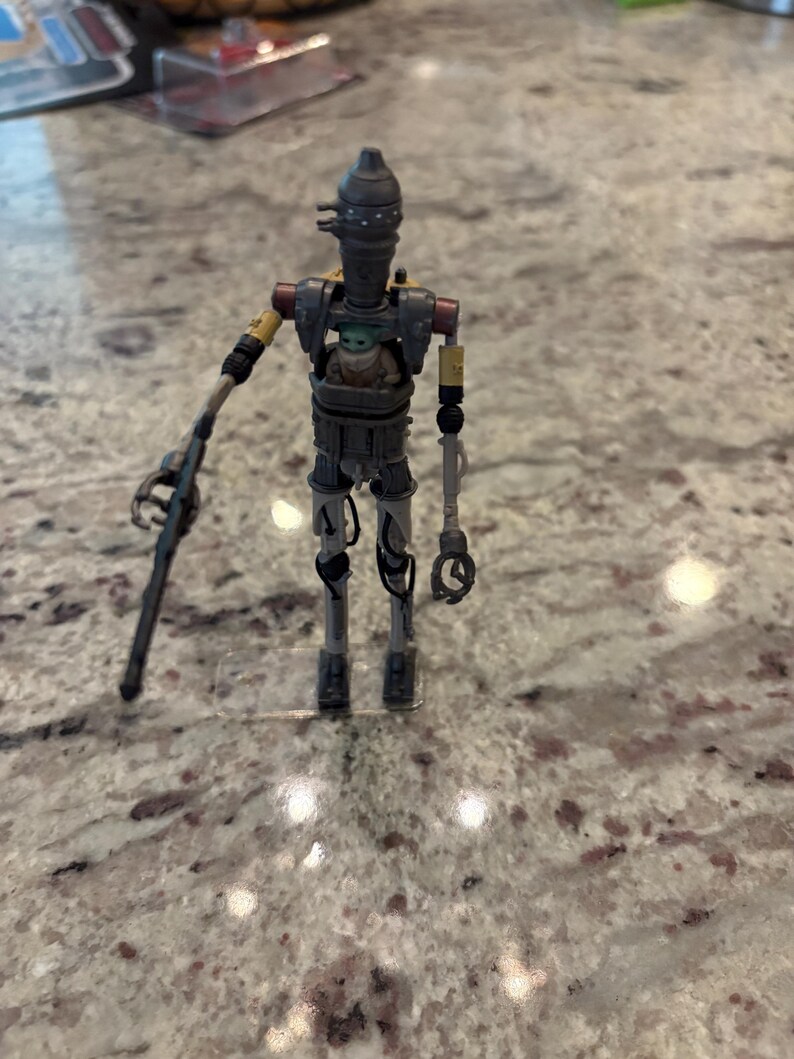 以下が含まれることがあります： スターウォーズのIG-11アクションフィギュア。中にはベビーヨーダのフィギュアが入っています。IG-11のフィギュアは主にグレーで、黄色と赤のアクセントがあります。ブラスターライフルを持っています。ベビーヨーダのフィギュアは緑と茶色です。