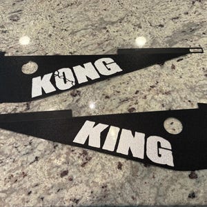 May include: Two black, triangular metal pieces with the words "KONG" and "KING" in white block letters. Each piece has a circular hole and a rectangular cutout. The items are displayed on a speckled gray surface.