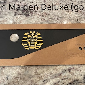 May include: A black and gold pinball machine side art featuring the Iron Maiden mascot, Eddie, in a pharaoh costume. The art is labeled "Iron Maiden Deluxe (gold)" and "Pinballers" with the text "extenders".