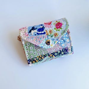 Könnte beinhalten: Eine kleine Geldbörse aus gestepptem Stoff mit einem Patchwork-Design aus verschiedenen Blumen- und Musterdrucken in Blau-, Rosa-, Grün- und Gelbtönen. Die Geldbörse hat einen dreieckigen Klappenverschluss mit einem silberfarbenen Metalldruckknopf.