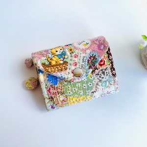 May include: A small fabric wallet featuring a patchwork design with kittens in baskets, floral patterns, and the text "LOVE" and "for you". The wallet has a snap closure and two round fabric-covered charms hanging from the side.