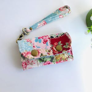 Könnte beinhalten: Eine kleine Patchwork-Geldbörse aus Stoff mit einem Handgelenkband. Die Geldbörse besteht aus verschiedenen floralen Stoffen in den Farben Rosa, Rot, Blau und Grün. Sie hat drei Messingknöpfe.