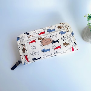 May include: Cream-colored zippered wallet featuring a repeating pattern of cartoon cats in various colors. The wallet has a small leather tag and a black zipper pull. The overall design is playful and suitable for cat lovers.