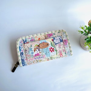 以下が含まれることがあります： 青、ピンク、クリーム色のパッチワークデザインのキルト財布。猫の愛らしいイラストと花の刺繍が特徴です。茶色の革のジッパープルと小さな革のタグが含まれています。