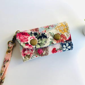 Könnte beinhalten: Eine kleine Patchwork-Geldbörse aus Stoff mit Blumenmuster und zwei Messingknöpfen. Die Geldbörse hat einen rosa und floral gemusterten Riemen.