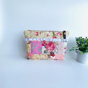 May include: A rectangular zipped pouch featuring a patchwork design of various floral and gingham fabrics in shades of pink, cream, and green. The top section is accented with a white lace trim, and a dark brown leather zip pull is attached.