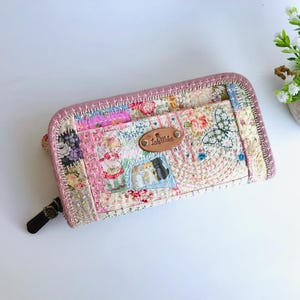 Könnte beinhalten: Eine handgefertigte Patchwork-Geldbörse mit Blumen- und Schmetterlingsdesign. Die Geldbörse hat eine Vielzahl von Stoffen in Rosa, Weiß und Blau, mit einem braunen Lederetikett. Die Kanten sind mit einer rosa Naht versehen.