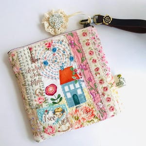 May include: Quilted fabric pouch with a floral and house design. The pouch has a zip closure, a brown leather wrist strap with a rose detail, and a decorative lace flower. The text "I see" and "Scatter See Happ" are embroidered on the pouch.