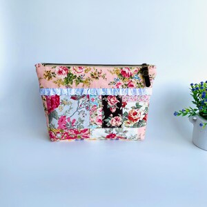 May include: A rectangular zipped pouch featuring a patchwork design of various floral fabrics in shades of pink, red, white, and black. The pouch is accented with a horizontal strip of white lace trim across the upper section and a dark zip pull.