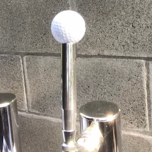 May include: A golf ball mounted on a polished metal stand. The white golf ball features a dimpled surface. The stand is cylindrical and has a shiny, reflective finish. The background is a grey, textured wall.