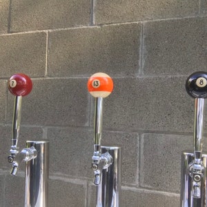 May include: Three beer tap handles with pool ball designs. The handles are red with the number 7, orange with the number 13, and black with the number 8. The taps are made of shiny metal and are mounted on a grey brick wall.