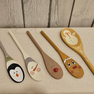May include: Five wooden spoons with painted holiday designs. The spoons feature a penguin, snowman, reindeer, and gingerbread person. The spoons are painted with various colors, including black, white, brown, and red. Each spoon has a long handle.