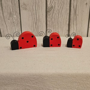 May include: Three wooden ladybug figurines, painted red with black spots and black heads. The ladybugs are arranged in a row on a white surface.