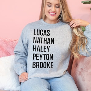 May include: A heather gray sweatshirt with the names Lucas, Nathan, Haley, Peyton, and Brooke printed in black, stacked vertically. The sweatshirt is a crew neck style.