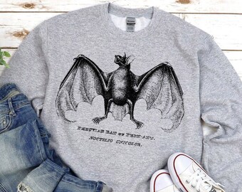 May include: A gray sweatshirt featuring a detailed black illustration of a bat with outstretched wings. The text "PERUVIAN BAT OF PERU, NOCTILIO UNICOLOR" is printed below the bat. The sweatshirt is paired with blue jeans and white sneakers.