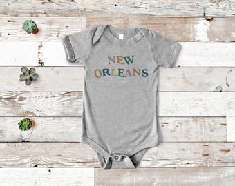 May include: A gray baby bodysuit with the words "NEW ORLEANS" in colorful, rounded letters. The bodysuit has short sleeves and a snap closure. The text is in shades of purple, green, blue, and gold.
