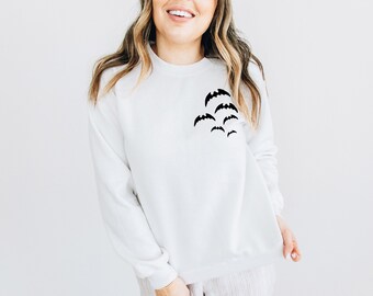 May include: White sweatshirt with a crew neck and long sleeves. A cluster of black bat silhouettes is printed on the left chest. The sweatshirt is a casual, comfortable garment, perfect for Halloween or everyday wear.