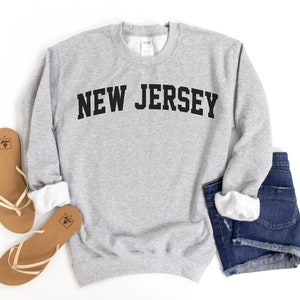 May include: A light gray sweatshirt with the text "NEW JERSEY" in bold, black letters. The sweatshirt is paired with denim shorts and brown sandals with white straps.