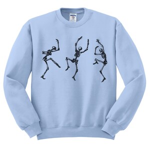 May include: Light blue sweatshirt featuring a black graphic of three dancing skeletons. The skeletons are in various poses, with arms and legs outstretched, giving a playful and spooky vibe. The sweatshirt is a crew neck style.
