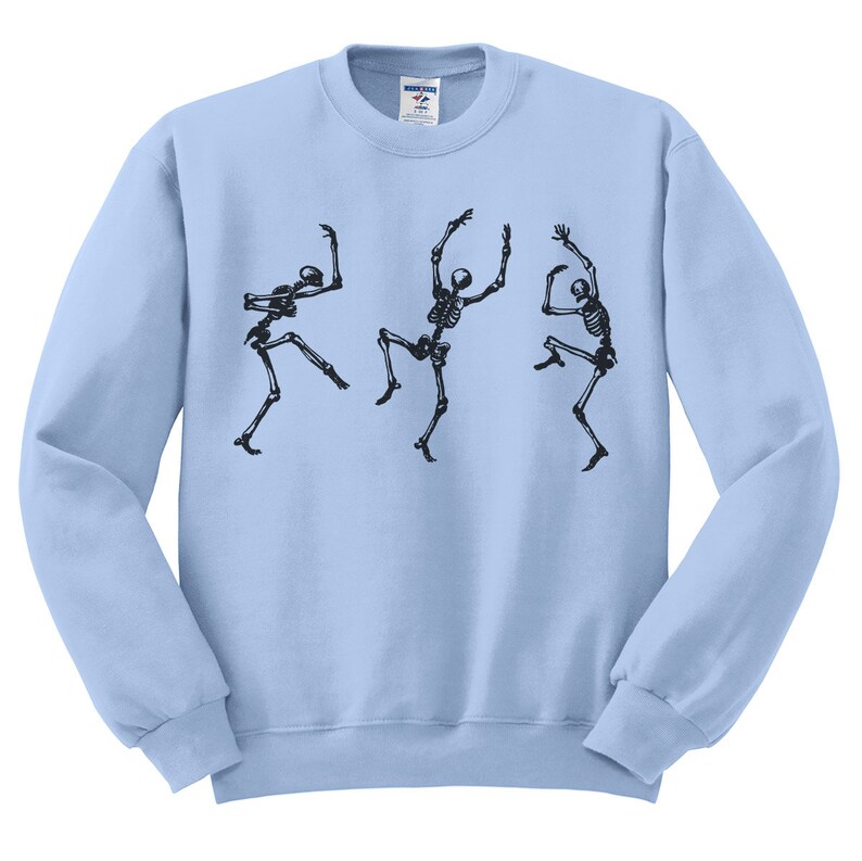 May include: Light blue sweatshirt featuring a black graphic of three dancing skeletons. The skeletons are in various poses, with arms and legs outstretched, giving a playful and spooky vibe. The sweatshirt is a crew neck style.