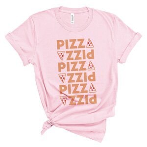 May include: A light pink t-shirt with rolled-up sleeves. The shirt features the word "PIZZA" stacked vertically five times in a brown color, with a pizza slice graphic next to each word. The shirt is knotted at the bottom.