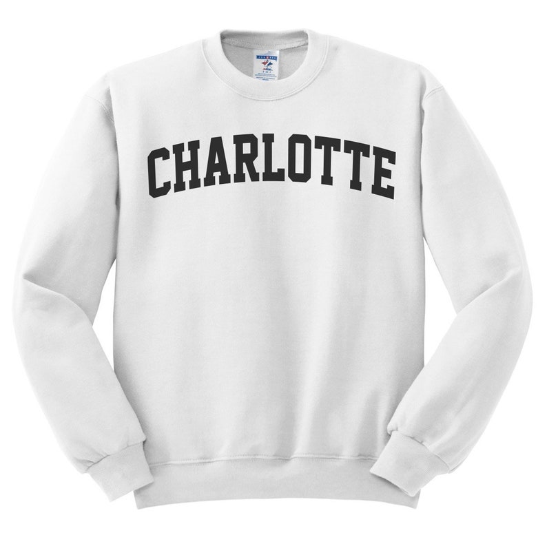 May include: White crew neck sweatshirt with the word "CHARLOTTE" printed across the chest in bold, black, arched letters. The sweatshirt has long sleeves and a ribbed collar, cuffs, and hem.