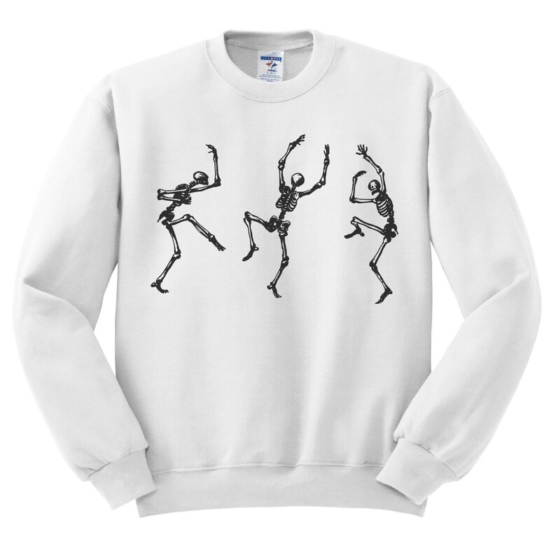 May include: White sweatshirt featuring a black graphic of three dancing skeletons. The skeletons are in various poses, with arms and legs outstretched, giving a playful, Halloween-themed design. The sweatshirt has a crew neck and long sleeves.