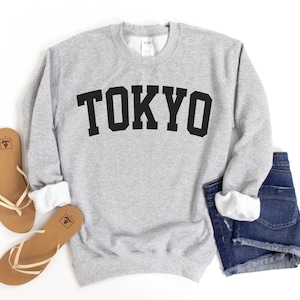 May include: A gray sweatshirt with the word "TOKYO" printed in black on the front. The sweatshirt is laid flat on a white background with a pair of brown sandals and denim shorts.