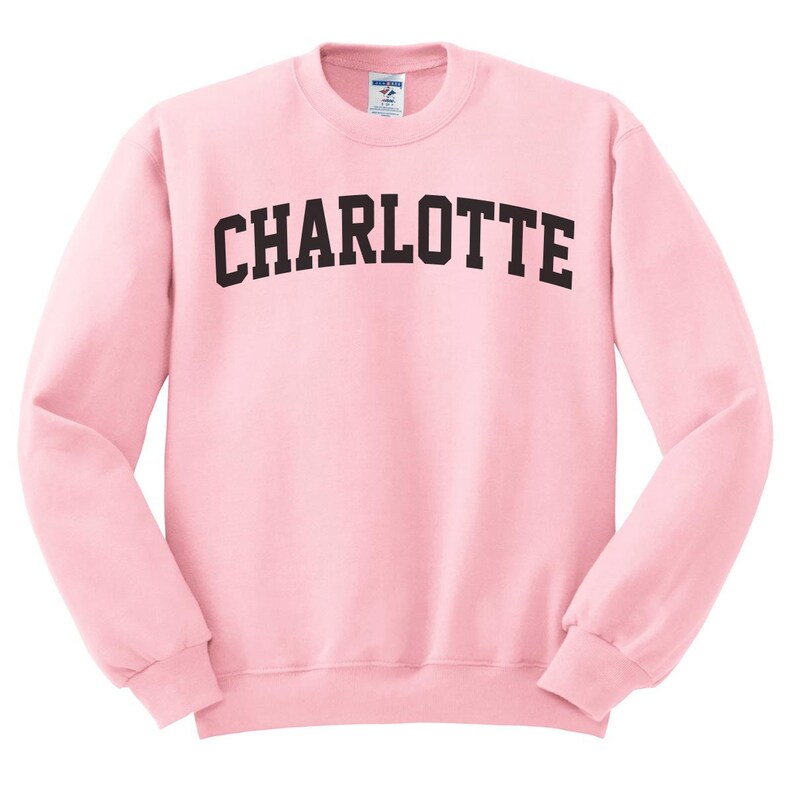 May include: A light pink crew neck sweatshirt with the word "CHARLOTTE" printed across the chest in bold, black, arched lettering. The sweatshirt has long sleeves and a ribbed hem and cuffs. The fabric appears soft and comfortable.