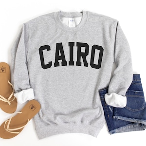 May include: Gray sweatshirt with black text that reads "CAIRO" in a collegiate font style. The sweatshirt is laid flat on a white surface with a pair of brown sandals and denim shorts.