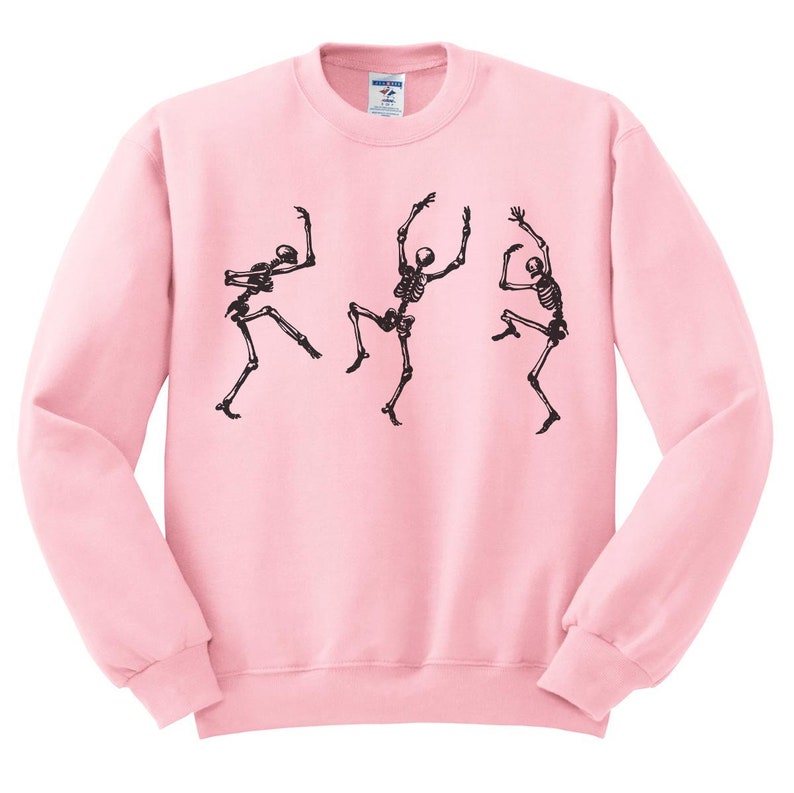May include: A light pink sweatshirt featuring a black graphic of three dancing skeletons. The skeletons are in various poses, with their arms and legs outstretched. The sweatshirt has a crew neck and long sleeves.