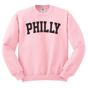 Puede incluir: Una sudadera rosa con la palabra "PHILLY" impresa en negro en la parte delantera.