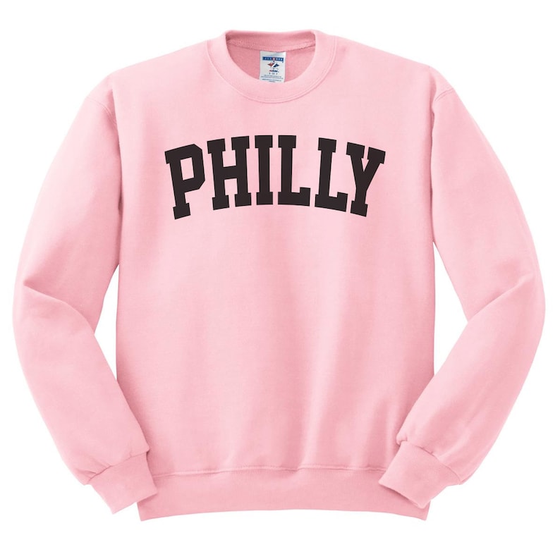 Puede incluir: Una sudadera rosa con la palabra "PHILLY" impresa en negro en la parte delantera.