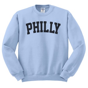 Puede incluir: Una sudadera de color azul claro con la palabra "PHILLY" impresa en negro en la parte delantera.